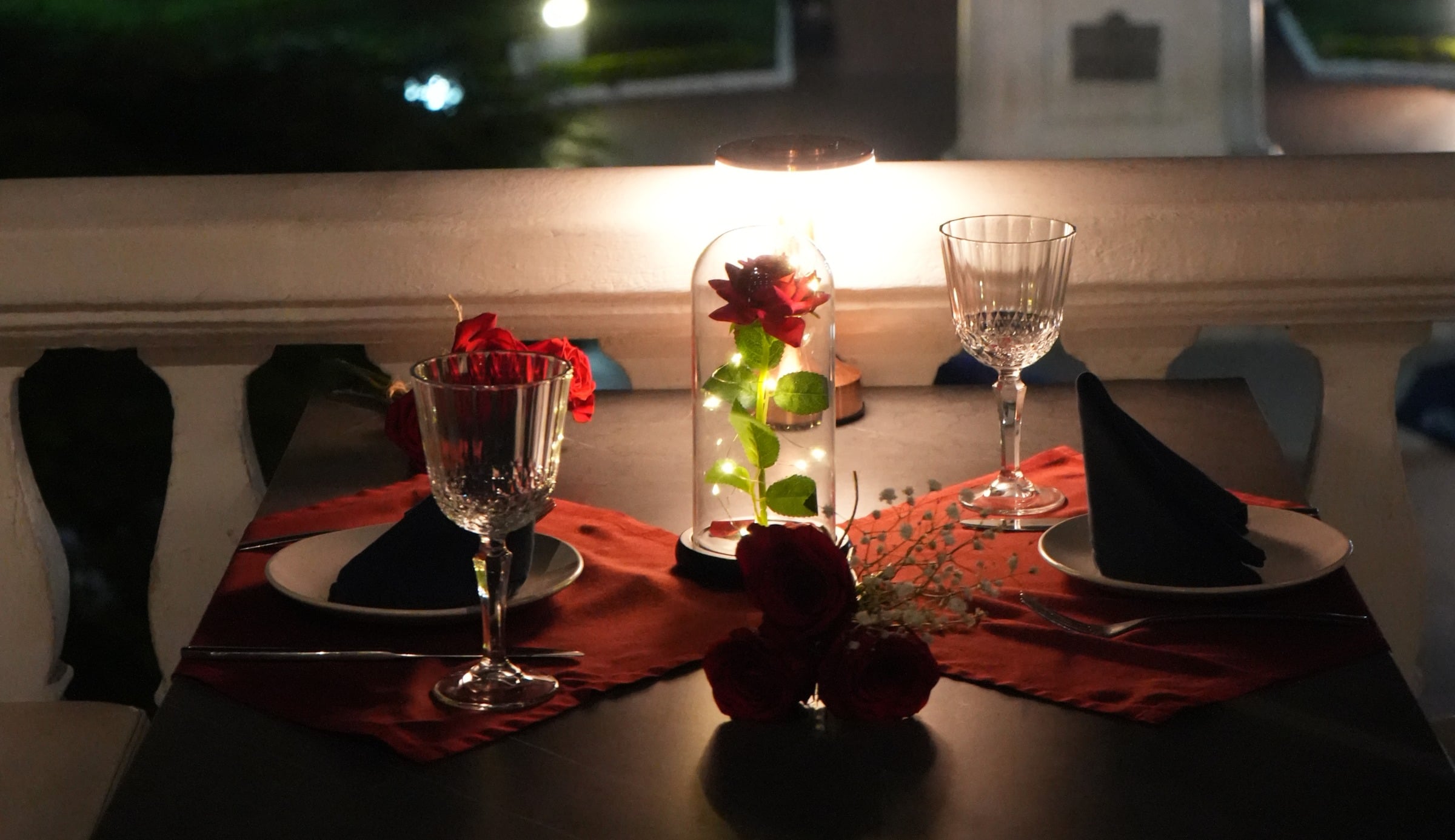 Mesa en el balcón con dos copas y una rosa bajo campana de cristal, frente a Plaza Herrera iluminada