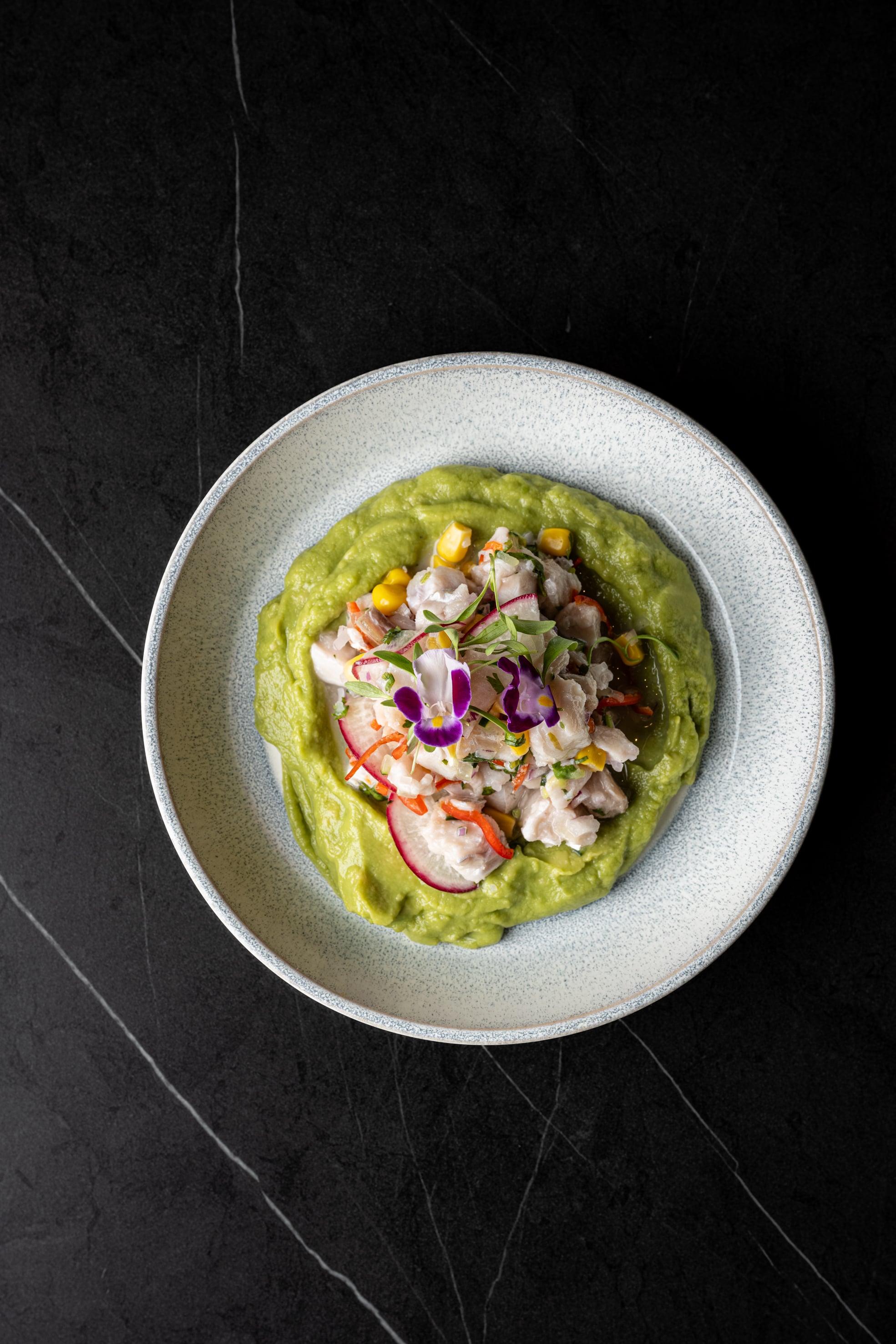 Ceviche sobre crema de aguacate con maíz, rábano y flores, en plato azul